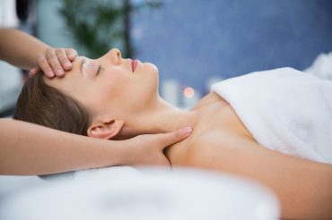 Profile portrait of young Caucasian woman lying on back, keeping her eyes closed while therapist massaging her head and shoulders. Spa concept